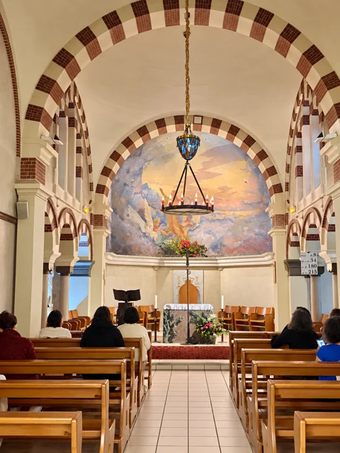Interior de uma capela com bancos e pintura atrás do altar.